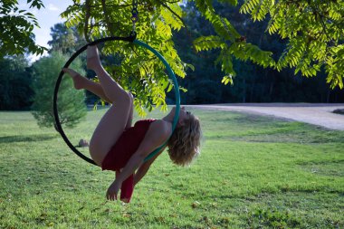 Kırmızı parti elbisesi giymiş genç sarışın bir kadın parkın ortasındaki ağaçtan sarkan bir halkayla egzersiz yapıyor. Kavramsal çember, direk dansı, fitness, kıvrımlı kız.