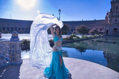 Orta yaşlı, turkuaz elbiseli, yapay elmaslı İspanyol kadın, beyaz peçeli göbek dansı. Göbek dansı konsepti.