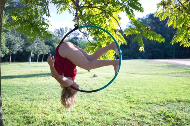 Kırmızı parti elbisesi giymiş genç sarışın bir kadın parkın ortasındaki ağaçtan sarkan bir halkayla egzersiz yapıyor. Kavramsal çember, direk dansı, fitness, kıvrımlı kız.