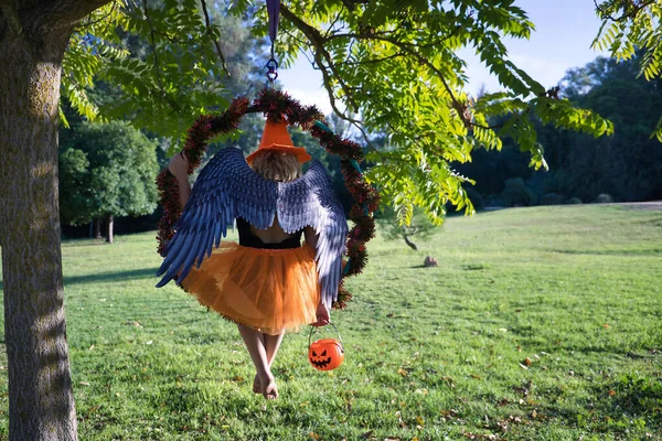 A young blonde woman in an orange hat and skirt with a pumpkin in her hand and with angel wings sitting on her back in an aerial ring that is hanging from a tree. Halloween concept, party, angels, costumes, aerial hoop, curvy girl.