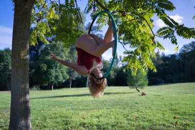 Kırmızı parti elbisesi giymiş genç sarışın bir kadın parkın ortasındaki ağaçtan sarkan bir halkayla egzersiz yapıyor. Kavramsal çember, direk dansı, fitness, kıvrımlı kız.