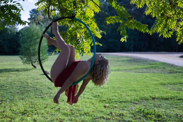 Young blonde woman in a red party dress exercising on an aerial hoop hanging from a tree in the middle of a park. Concept aerial hoop, pole dance, fitness, curvy girl.