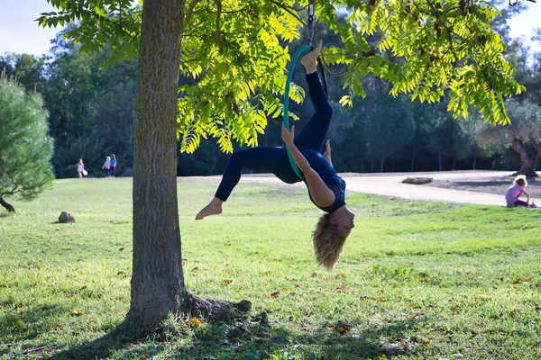 Young blonde woman in a blue suit, exercising upside down in an aero hoop hanging from a tree in the middle of a park. Concept aerial hoop, pole dance, fitness, curvy girl.