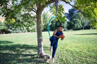 Cadı şapkası takan ve elinde bir balkabağı tutan genç sarışın bir kadın ağaçtan sarkan bir halkayla. Cadılar Bayramı konsepti, parti, sonbahar, hava çemberi, kıvrımlı kız.