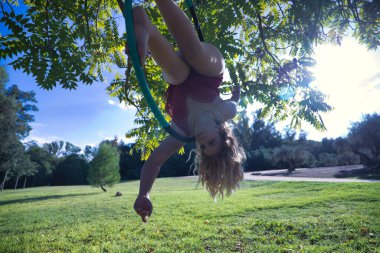 Kırmızı parti elbisesi giymiş genç sarışın bir kadın parkın ortasındaki ağaçtan sarkan bir halkayla egzersiz yapıyor. Kavramsal çember, direk dansı, fitness, kıvrımlı kız.