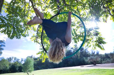 Mavi takım elbiseli genç sarışın bir kadın, parkın ortasındaki bir ağaçtan sarkan bir aero çemberin içinde baş aşağı egzersiz yaparken, güneş ışınlarını alıyor. Kavramsal çember, direk dansı, fitness.