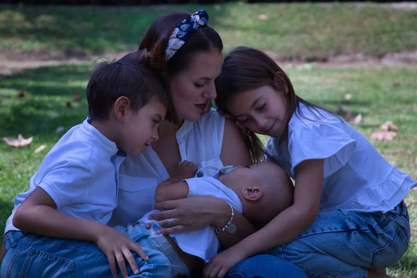 彼女の腕の中で眠っている赤ん坊と彼女の2人の子供と母は 屋外公園の芝生の上に座って 弟を見ています 概念的な母親 優しさ — ストック写真