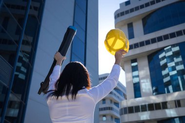 Beyaz gömlekli genç bir kadın, kolları açık, sarı işçi miğferi ve elinde plan borusuyla iyi bir işi kutluyor. İş konsepti, mimari, inşaat, güçlendirme.