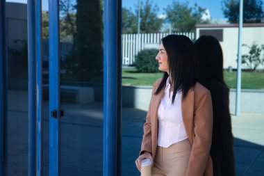 Kahverengi paltolu ve elinde bir bardak kahve olan genç, güzel bir kadın kahvaltı saatinde ofisinin kapısına yaslanıyor. İş konsepti, çalışan kadın, dinlenme, güçlendirme.