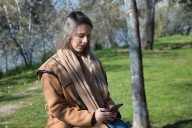 Genç sarışın kadın, bir bankın tepesinde oturuyor, parkta, cep telefonundaki sosyal ağlara danışıyor. Akıllı telefonu, sosyal ağları, bağlantıyı, bağımlılığı kavra.