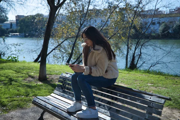 Молодая Брюнетка Очках Сидит Скамейке Парке Проверяет Социальные Сети Мобильном — стоковое фото
