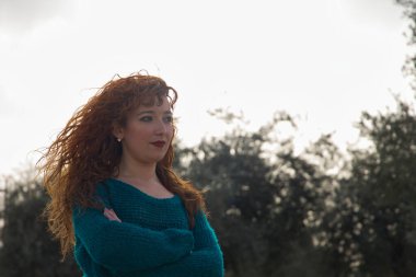 Kızıl saçlı, çilli, kollarını kavuşturmuş ve açık hava parkında ciddi jestler yapan genç bir kadın. Yalnızlığı, ciddiyeti, kederi, tasayı kavra..