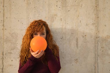 Genç, çekici, kızıl saçlı, çilli, kırmızı kazaklı genç bir kadın gri bir duvarda turuncu bir balonu dolduruyor. Kavram renkleri, balonlar, güzellik, moda, kırmızı.