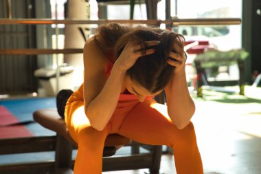 Spor salonundaki bankta oturan genç ve güzel bir kadın elleri başının üstünde, yorgun ve depresif. Depresyon, yorgunluk, stres, spor, yalnızlık, aşırı eğitim.