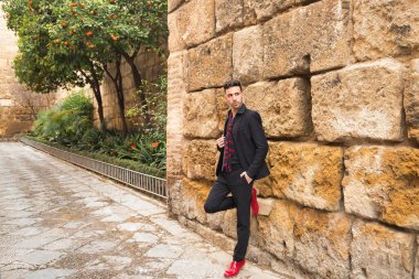 Siyah gömlek, ceket ve pantolon giyen, kırmızı puantiyeli siyah atkı giyen, kırmızı dans ayakkabısı giyen, taş bir duvarın yanında poz veren genç bir İspanyol. Flamenko konsepti, sanat, dans, kültür, gelenek.