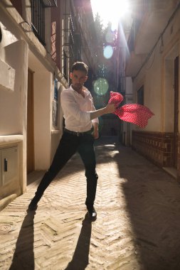 Beyaz tişörtlü, siyah pantolonlu ve ayakkabılı genç İspanyol adam, kırmızı mendilli flamenko dans ediyor. Sokakta, güneş ışınlarının altında beyaz benekli. Kavram dansı, kültür, gelenek.