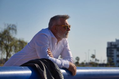 Olgun, gri saçlı, sakallı, güneş gözlüklü, beyaz gömlekli, deri ceketli, ufka endişeli bir şekilde bakan bir adam. Olgun erkek kavramı, emeklilik, işsizlik, sorunlar, yalnızlık.