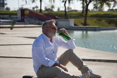 Olgun, sakallı, güneş gözlüklü, beyaz gömlekli, açık kahverengi pantolonlu, ağzında bir şişe birayla gölün yanında oturan bir adam. Olgun erkek kavramı, alkol, içki, tatil, sorunlar.