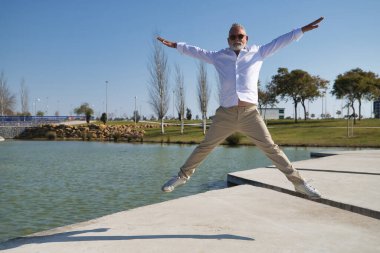 Olgun bir adam, gri saçlı, sakallı, güneş gözlüklü, beyaz gömlekli ve açık kahverengi pantolonlu, göl kenarında mutlu bir şekilde havaya atlıyor. Mutluluk kavramı, neşe, atlama, tatil, emeklilik.