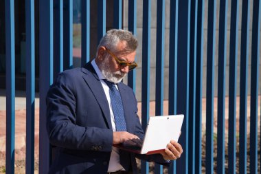 Olgun bir adam, yönetici, gri saçlı, sakallı, güneş gözlüklü, ceketli ve kravatlı, ofisin çıkışındaki laptoptan e-postaları kontrol eden. Yönetici kavramı, iş adamı, iş adamı, uygulamalar, dizüstü bilgisayar.