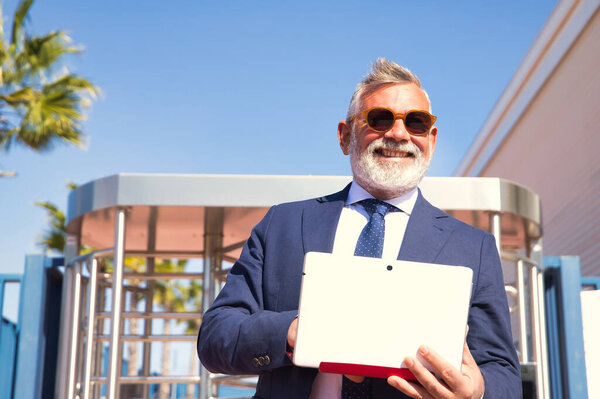 Mature man, rector, gray-haired, bearded, sunglasses, jacket and tie, consulted happily, emails on laptop at the exit of the university. Professor concept, businessman, business, applications, laptop.