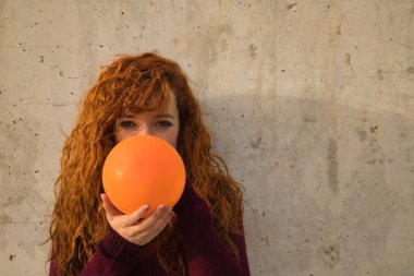 Gri bir duvarda turuncu bir küreyi dolduran genç, çekici, kızıl saçlı, çilli, kırmızı kazaklı bir kadının portresi. Kavram renkleri, balonlar, güzellik, moda, kırmızı.