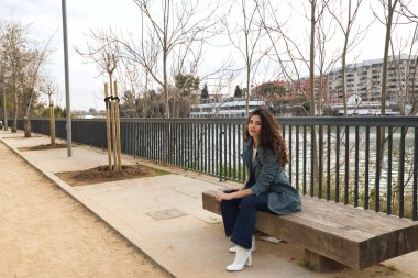 Genç ve güzel bir kadın, esmer, kıvırcık saçlı, ceketli, kot pantolonlu ve beyaz çizmeli, tahta bir bankta oturuyor. Güzellik, moda, trend, model, sonbahar kavramları.