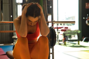 Spor salonundaki bankta oturan genç ve güzel bir kadın elleri başının üstünde, yorgun ve depresif. Depresyon, yorgunluk, stres, spor, yalnızlık, aşırı eğitim.