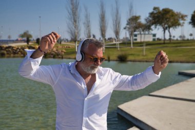 Olgun, sakallı, güneş gözlüklü, beyaz gömlekli, beyaz kulaklıklı, göl kenarında müzik dinlerken mutlu bir şekilde dans eden bir adam. Mutluluk kavramı, neşe, müzik, disko, dans, emeklilik.