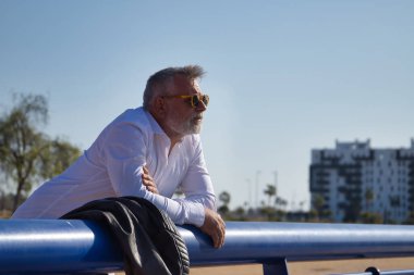 Olgun, gri saçlı, sakallı, güneş gözlüklü, beyaz gömlekli, deri ceketli, ufka endişeli bir şekilde bakan bir adam. Olgun erkek kavramı, emeklilik, işsizlik, sorunlar, yalnızlık.