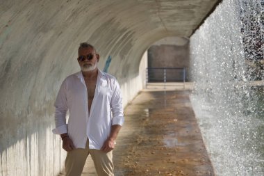 Olgun, gri saçlı, sakallı, güneş gözlüklü, beyaz gömlekli, açık kahverengi pantolonlu, şelalenin yanındaki beton tünelin altında. Olgun bir adam, zengin bir baba, yaşam tarzı, oyuncak ayı.