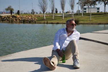 Olgun, gri saçlı, sakallı, güneş gözlüklü, beyaz gömlekli, açık kahverengi pantolonlu, elinde bir şişe birayla göl kenarında oturan bir adam. Olgun erkek kavramı, alkol, içki, tatil, sorunlar.