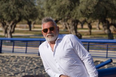 Olgun bir adam, gri saçlı, sakallı, güneş gözlüklü, beyaz gömlekli, dışarıda korkuluklara yaslanmış. Büyükbaba, zengin baba, yaşam tarzı, olgun adam.