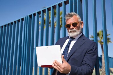 Olgun bir adam, profesör, gri saçlı, sakallı, güneş gözlüklü, ceketli ve kravatlı, üniversitenin çıkışında e-posta kutusuna bakıyor. Rektör konsepti, iş adamı, iş adamı, uygulamalar, dizüstü bilgisayar.