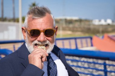 Olgun bir adam, yönetici, kır saçlı, sakallı, güneş gözlüklü, ceketli ve kravatlı, dişleriyle kriptoyu ısırıyor. Yönetici kavramı, bitcoin, business, mines.