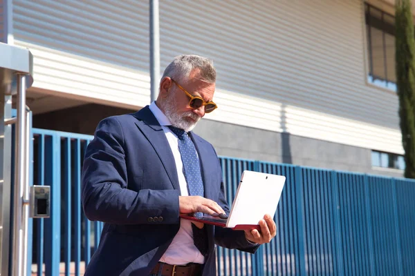 Olgun bir adam, yönetici, gri saçlı, sakallı, güneş gözlüklü, ceketli ve kravatlı, ofisin çıkışındaki laptoptan e-postaları kontrol eden. Yönetici kavramı, iş adamı, iş adamı, uygulamalar, dizüstü bilgisayar.