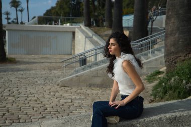 Genç ve güzel bir kadın, esmer, kıvırcık saçlı, kayıp bakışlı, beyaz gömlek ve kot pantolon giyen, beton bir duvarda oturan. Güzellik, moda, endişe, sorunlar, yalnızlık kavramı.