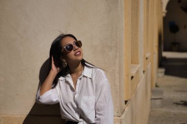 Beyaz gömlekli, kot pantolonlu ve güneş gözlüklü güzel bir Güney Amerikalı kadının portresi Avrupa 'da tatilde bir duvarda güneşleniyor, saçlarına dokunuyor. Güzellik, moda, tatil, seyahat kavramı.