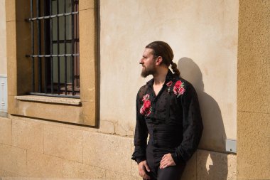 Sakallı, at kuyruklu, flamenko dansçısı, siyah şeffaf gömlekli, puantiyeli, kırmızı güllü, siyah pantolonlu ve ceketli, duvara yaslanmış genç bir adam. Sanat, dans, kültür kavramı.