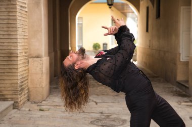 Sakallı, uzun saçlı, siyah şeffaf gömlekli, siyah puantiyeli, kırmızı güllü, siyah pantolonlu ve ceketli, şehirde flamenko dansı yapan genç bir adam. Sanat, dans, kültür, gelenek.