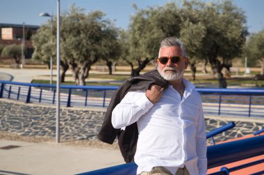 Mature, gray haired, bearded man with sunglasses, white shirt and leather jacket over his shoulder, leaning on a railing in the open air. Concept grandfather, sugar daddy, lifestyle, mature man.