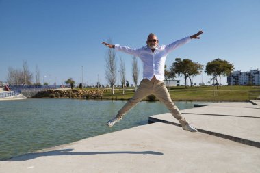 Olgun bir adam, gri saçlı, sakallı, güneş gözlüklü, beyaz gömlekli ve açık kahverengi pantolonlu, göl kenarında mutlu bir şekilde havaya atlıyor. Mutluluk kavramı, neşe, atlama, tatil, emeklilik.