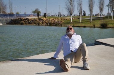 Olgun, gri saçlı, sakallı, güneş gözlüklü, beyaz gömlekli, açık kahverengi pantolonlu, gölün kenarında yatan bir adam. Olgun erkek kavramı, emeklilik, işsizlik, tatil, yalnızlık.