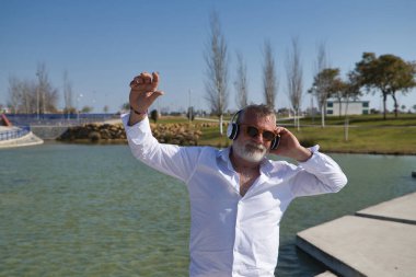 Olgun, sakallı, güneş gözlüklü, beyaz gömlekli, beyaz kulaklıklı, göl kenarında müzik dinlerken mutlu bir şekilde dans eden bir adam. Mutluluk kavramı, neşe, müzik, disko, dans, emeklilik.