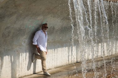 Olgun, gri saçlı, sakallı, güneş gözlüklü, beyaz gömlekli ve açık kahverengi pantolonlu, bir şelale altında beton duvara yaslanmış. Olgun bir adam, zengin bir baba, yaşam tarzı, oyuncak ayı.