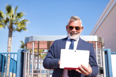 Olgun bir adam, rektör, gri saçlı, sakallı, güneş gözlüğü, ceket ve kravat takan, mutlu bir şekilde danışan, üniversitenin çıkışında dizüstü bilgisayardaki e-postalar. Profesör konsepti, iş adamı, iş adamı, başvuru, dizüstü bilgisayar.