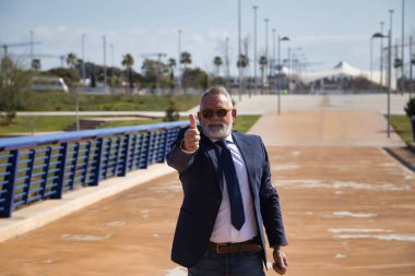 Olgun bir adam, yönetici, gri saçlı, sakallı, güneş gözlüklü, ceketli ve kravatlı, parmakları havada, mutlu, çünkü işler iyi gidiyor. Kavram yöneticisi, işadamı, iş adamı, istihdam, zafer.