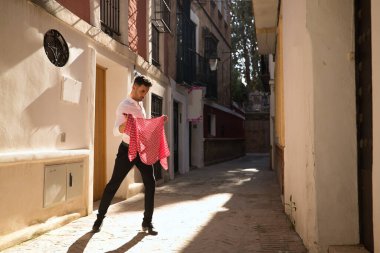 Beyaz gömlekli, siyah pantolonlu, dans ayakkabılı, kırmızı mendilli, dar bir sokakta beyaz benekli bir flamenko dans eden genç bir İspanyol. Sanat, dans, kültür, gelenek.