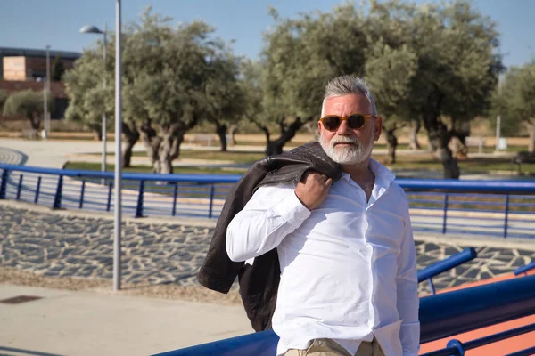 Olgun, gri saçlı, güneş gözlüklü, beyaz gömlekli, deri ceketli, açık havada korkuluklara yaslanmış bir adam. Büyükbaba, zengin baba, yaşam tarzı, olgun adam.