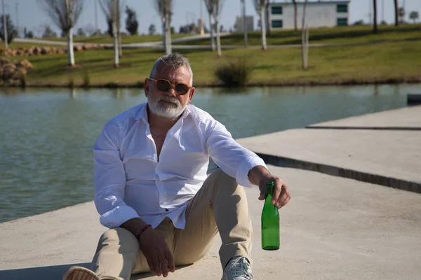 Olgun, gri saçlı, sakallı, güneş gözlüklü, beyaz gömlekli, açık kahverengi pantolonlu, elinde bir şişe birayla göl kenarında oturan bir adam. Olgun erkek kavramı, alkol, içki, tatil, sorunlar.